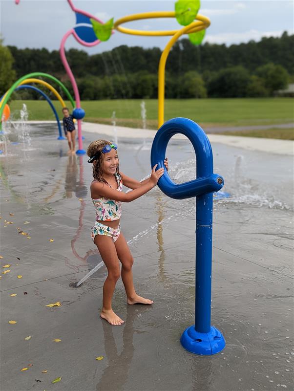 A child is playing with a blue water feature at a splash park, with colorful structures in the background and another child visible.