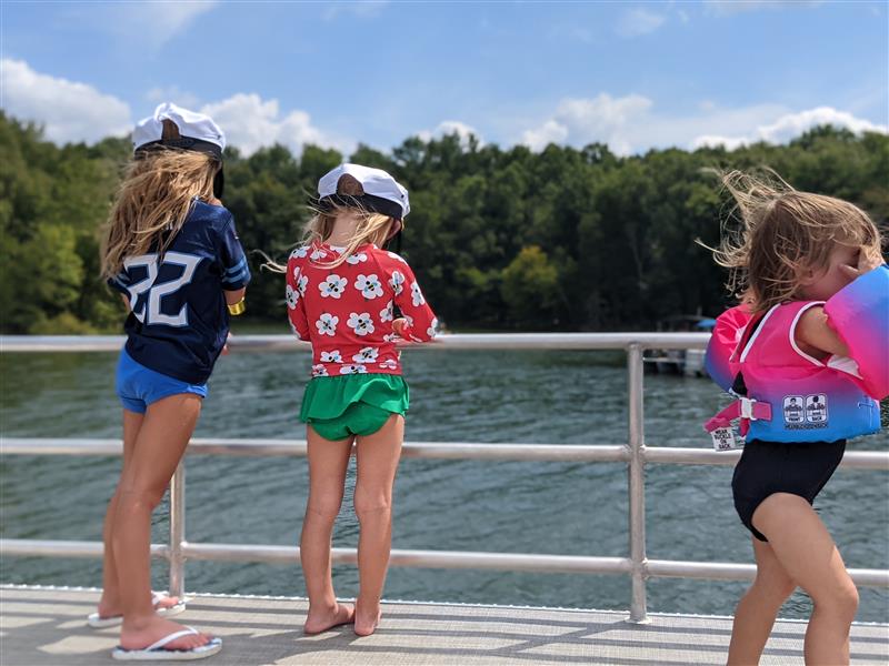 Three children stand on a dock overlooking a body of water with forest in the background. They wear swimwear and sports apparel, two with hats.