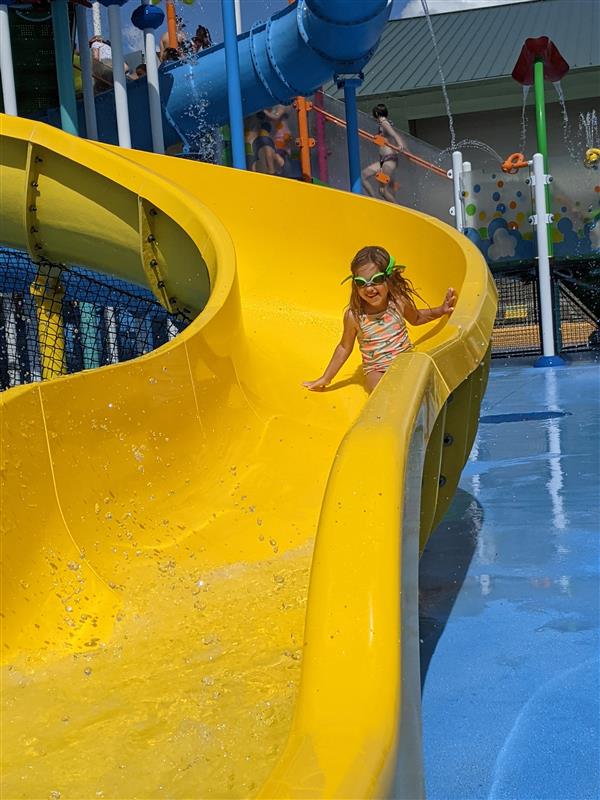 A child is sliding down a bright yellow water slide at a colorful water park, lined with splashing water features and water-soaked grounds.