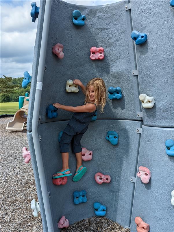 A child climbs on an outdoor artificial rock wall with colorful handholds, looking towards the camera with a smile, in a playground setting.