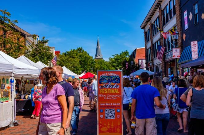 A lively street festival with people browsing vendor tents under a clear sky. Colorful banners and a church spire rise in the background.