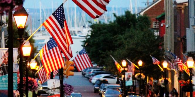 Street lined with American flags, illuminated by street lamps at dusk, with parked cars and a harbor in the background, evoking a patriotic atmosphere.