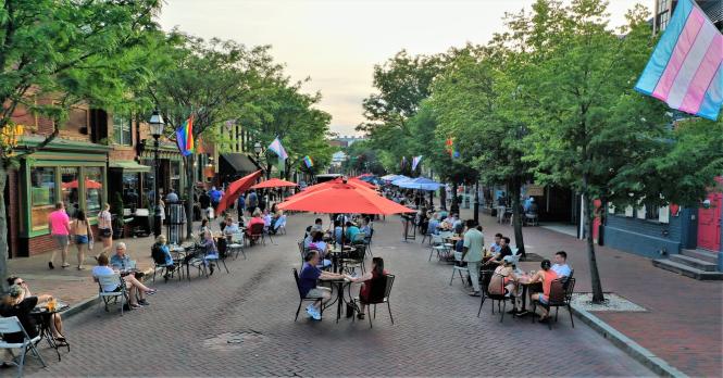 This image shows a lively street scene with people dining outdoors, colorful flags fluttering above, and a brick-lined pedestrian area flanked by leafy trees.