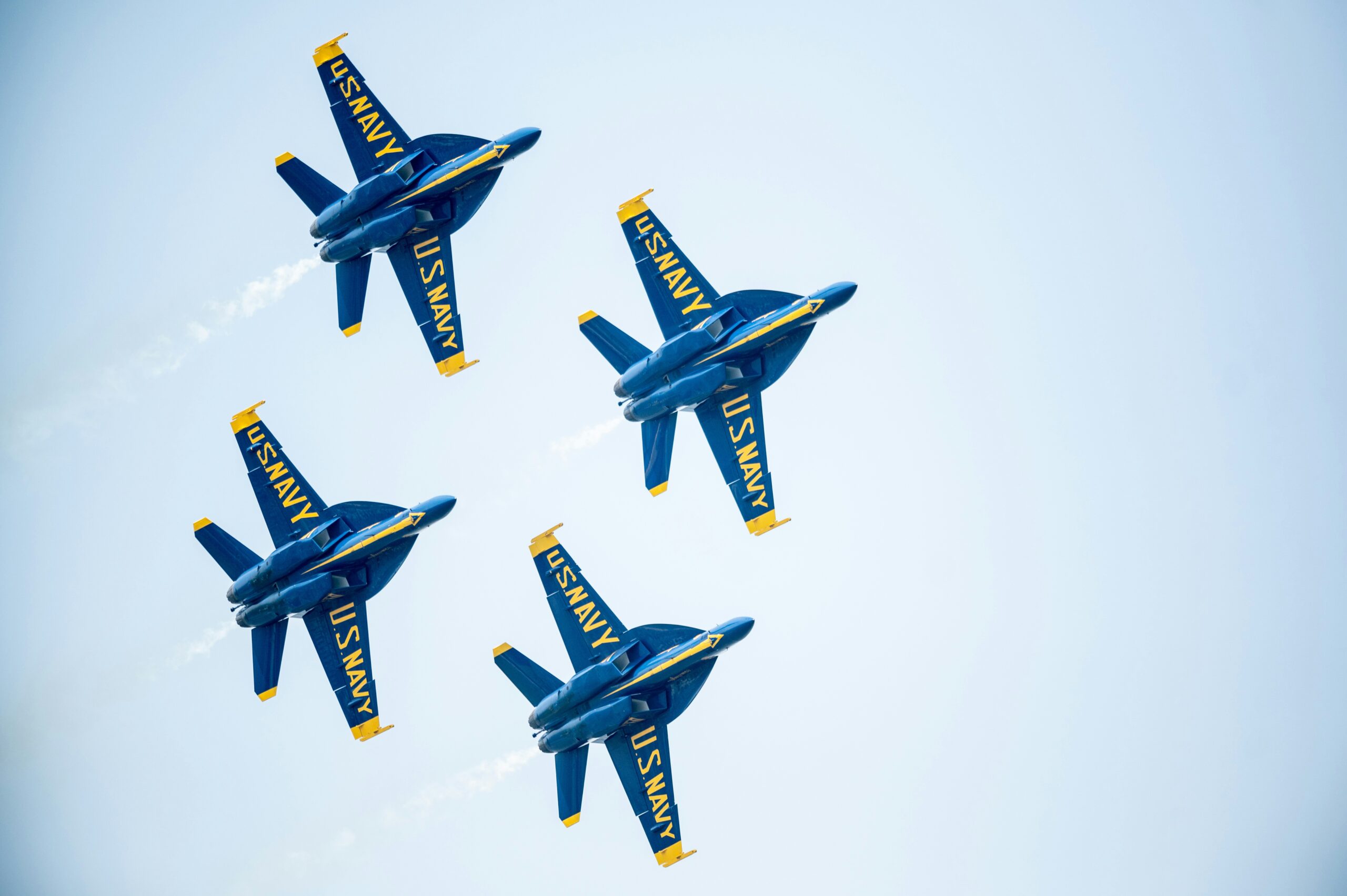 Five Blue Angels jets, with "US NAVY" markings, are flying in tight formation against a light blue sky, demonstrating precision and teamwork.