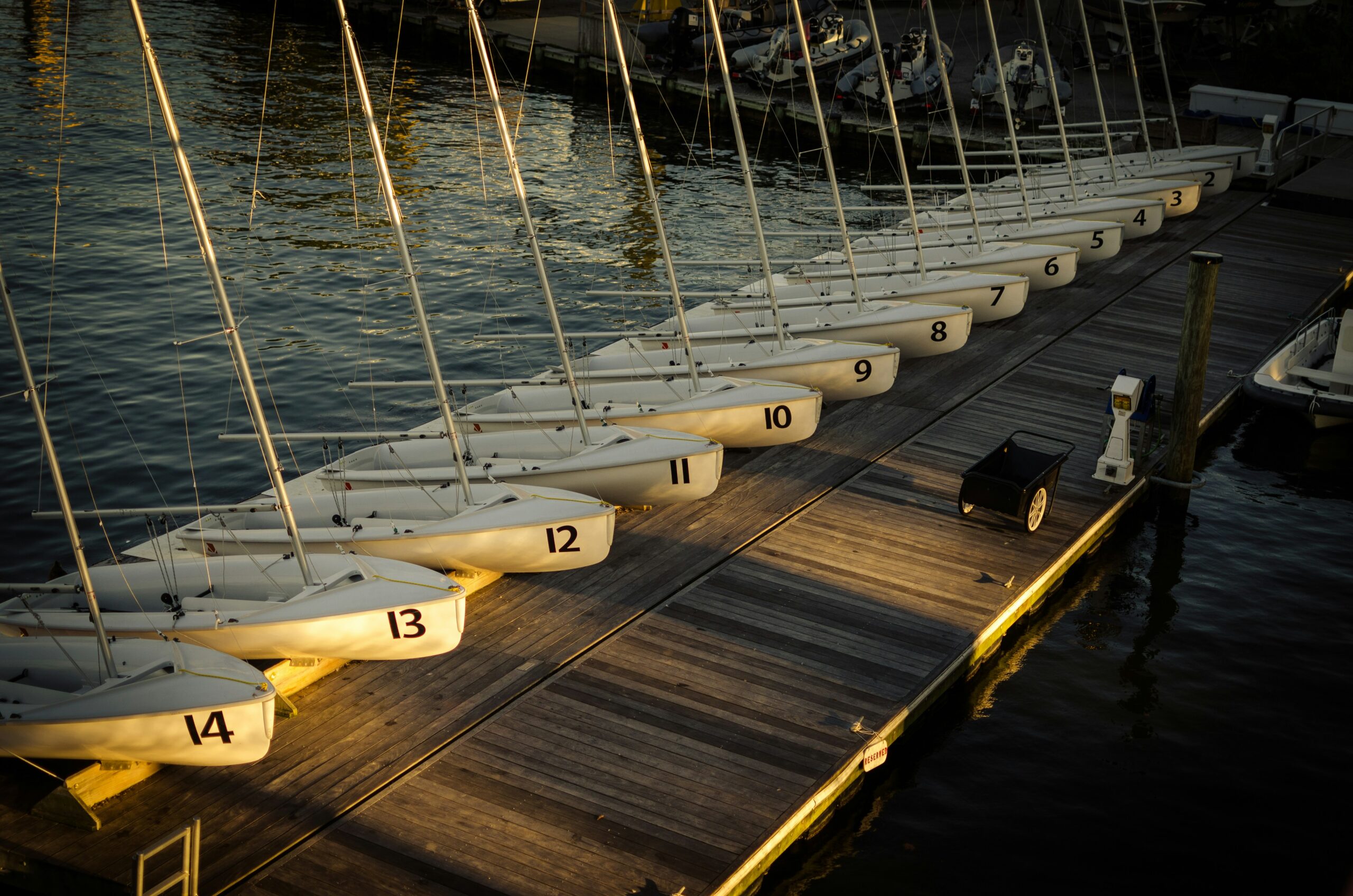 A row of small, white sailboats, numbered 5 through 14, docked at a wooden pier bathed in warm sunset light with calm waters surrounding them.