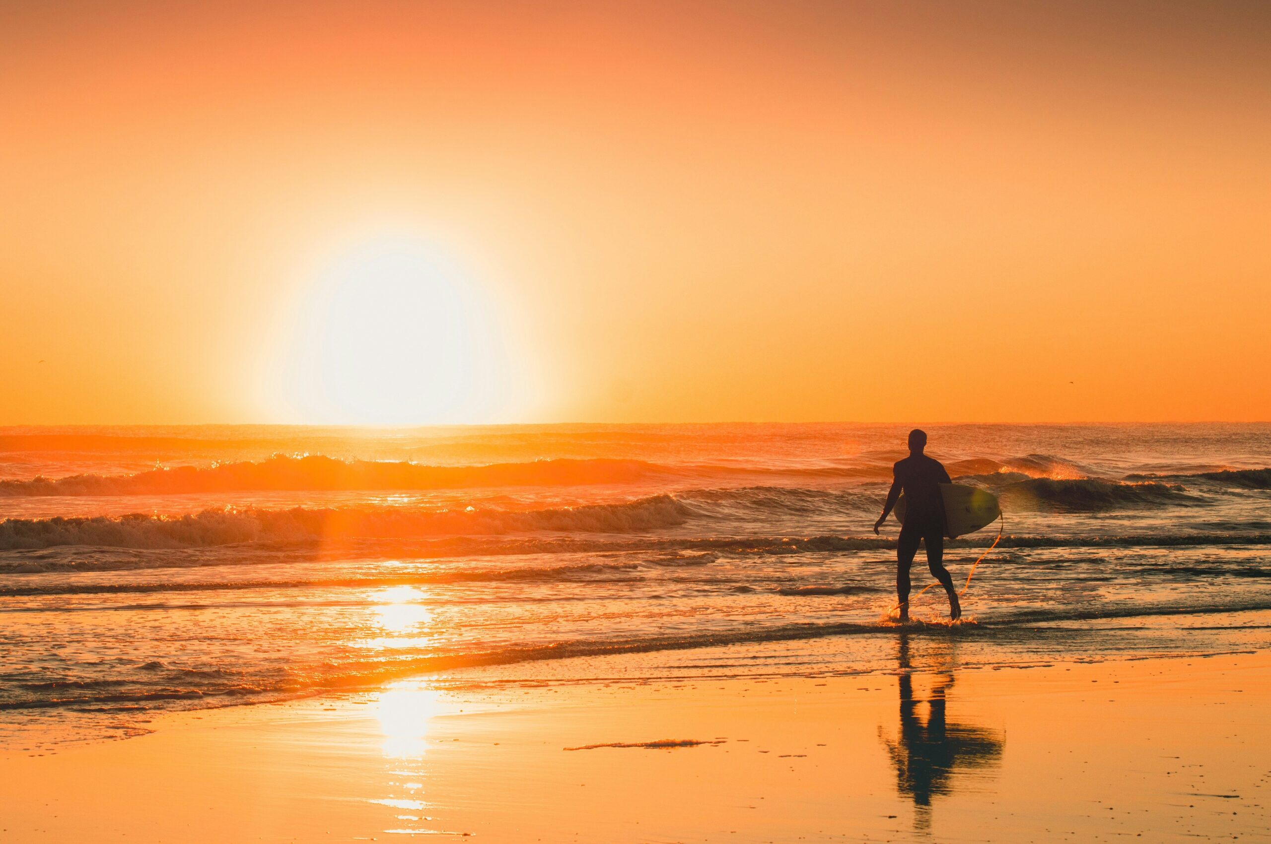 A person holding a surfboard walks along the beach at sunset, with waves gently breaking and the sky glowing warmly from the low sun.